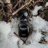 black gradient perfume bottle in the snow, twigs, and moss with two rusty coffin nails placed below. Bottle reads "HoloGrave Cosmetics", "eau de veil", and has a coffin tag that reads "Grave Mistakes"